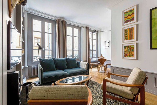 A modern living room with a dark green sofa, a wooden armchair, and a glass coffee table. Large windows provide natural light, and artwork is displayed on the walls. - Curiosité - Furnished apartment - Downtown Lyon (Lyon-1er-arrondissement)
