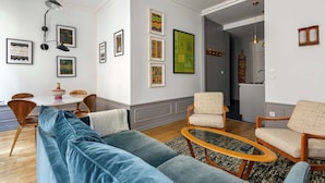 A modern living room with a blue sofa, a dining area with a table and chairs, and a kitchen visible through an open doorway.