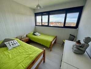 A small, tidy room with two single beds, a desk, a chair, a window with a view of buildings, and a statue on the desk.