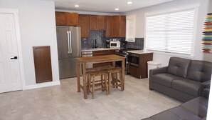 A compact kitchen with wooden cabinets, stainless steel appliances, a wooden table with stools, and a gray sofa.