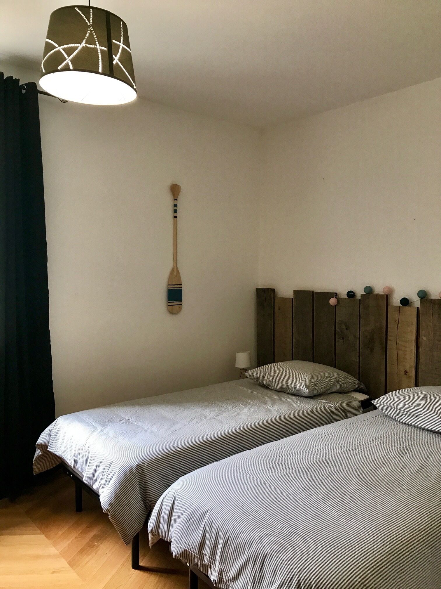 A room with two beds, a wooden headboard, a wall-mounted paddle, and a hanging light fixture.