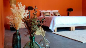 Een slaapkamer met een oranje wand, een bed met wit beddengoed, twee nachtkastjes met lampen, en drie vazen met gedroogde bloemen op de vloer.