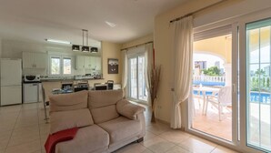 Een ruime woonkamer met een bank, een keuken op de achtergrond en een glazen schuifdeur naar een buiten zitgedeelte.