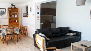 A living room with a black sofa, wooden dining table, chairs, and a bookshelf. - Gîte Écureuil: Lower Villa 2 Bedrooms, Terrace, Parking (Velaux)