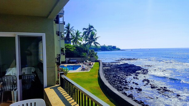 A balcony with a view of the ocean, a pool area, and a grassy lawn.