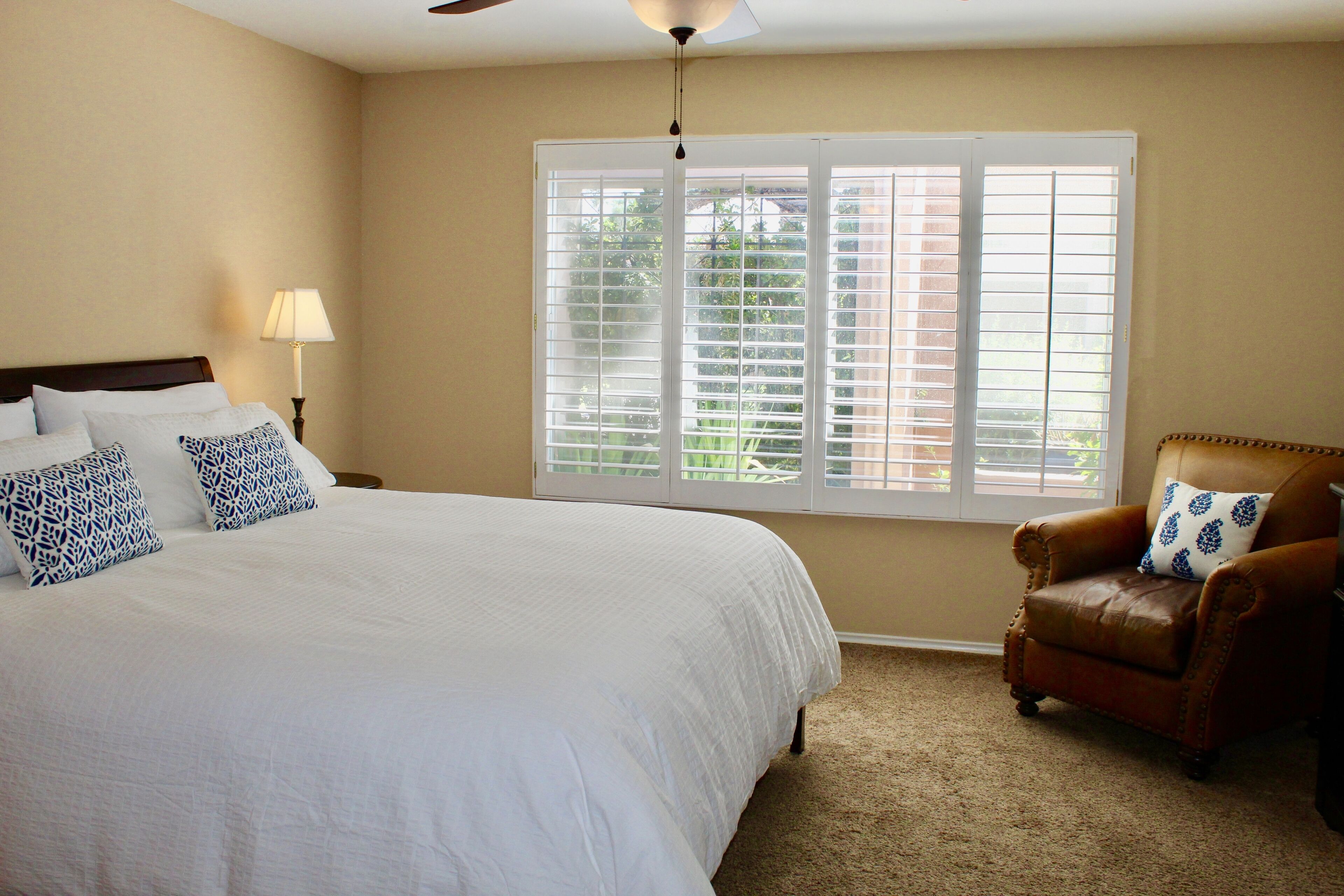 A bedroom with a large bed, a chair, a window with blinds, and a ceiling fan.