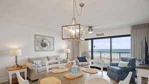 Ein modernes Wohnzimmer mit einem runden Holztisch, weißen Stühlen, einem blauen Sessel und Blick auf den Strand durch große Fenster.