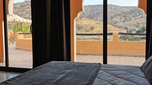 Una camera da letto con un letto, vista su un cortile con una sedia e piante, e un paesaggio montano all'esterno.