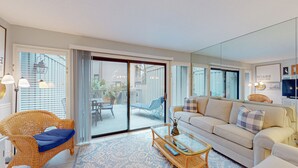Un salón espacioso con un amplio ventanal corredera que da a un patio exterior, un conjunto de sofás cómodos, una silla de mimbre y una mesa de centro de vidrio.