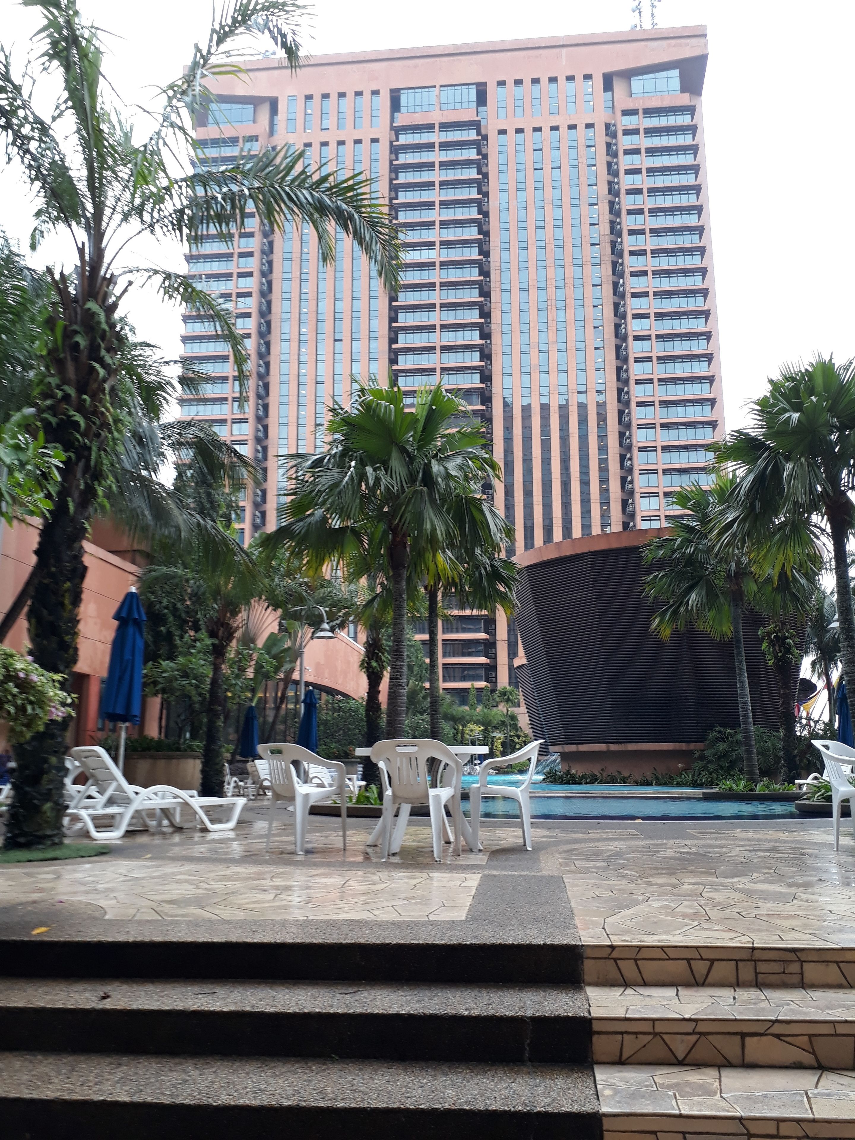 Un bâtiment moderne doté d’une façade réfléchissante, entouré de palmiers et comprenant un espace piscine avec des chaises longues.