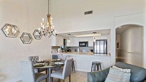 Une cuisine moderne avec des armoires blanches, des appareils en acier inoxydable et un îlot central. On y trouve un coin repas avec une table ronde et des chaises, ainsi qu’un canapé en cuir agrémenté d’un coussin décoratif à motifs.