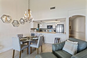 A modern kitchen with white cabinets, stainless steel appliances, and a central island. There is a dining area with a round table and chairs, and a leather sofa with a patterned throw pillow.
