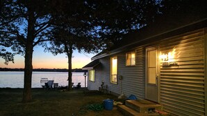A lakeside cabin with a dock, outdoor seating, and a well-lit entrance. - Hamptons style Lake house on Sunset point. With water on both the east and west! (Lindstrom)