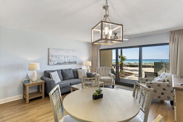 A modern living room with a round table, two sofas, and a dining area. There is a large window offering a view of the beach.