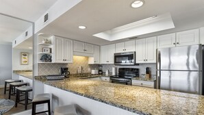 Cocina moderna con gabinetes blancos, encimeras de granito, electrodomésticos de acero inoxidable y una barra de desayuno con taburetes.