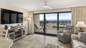 Un soggiorno spazioso con una TV a schermo piatto, un ventilatore a soffitto e un'ampia porta scorrevole in vetro che dà su un balcone con vista sull'oceano.