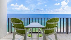 En balkon med to grønne Adirondack-stole og et lille bord, med udsigt over et klart, blåt hav og en strand.