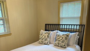 A bedroom with a bed, wooden flooring, and two windows with blinds.
