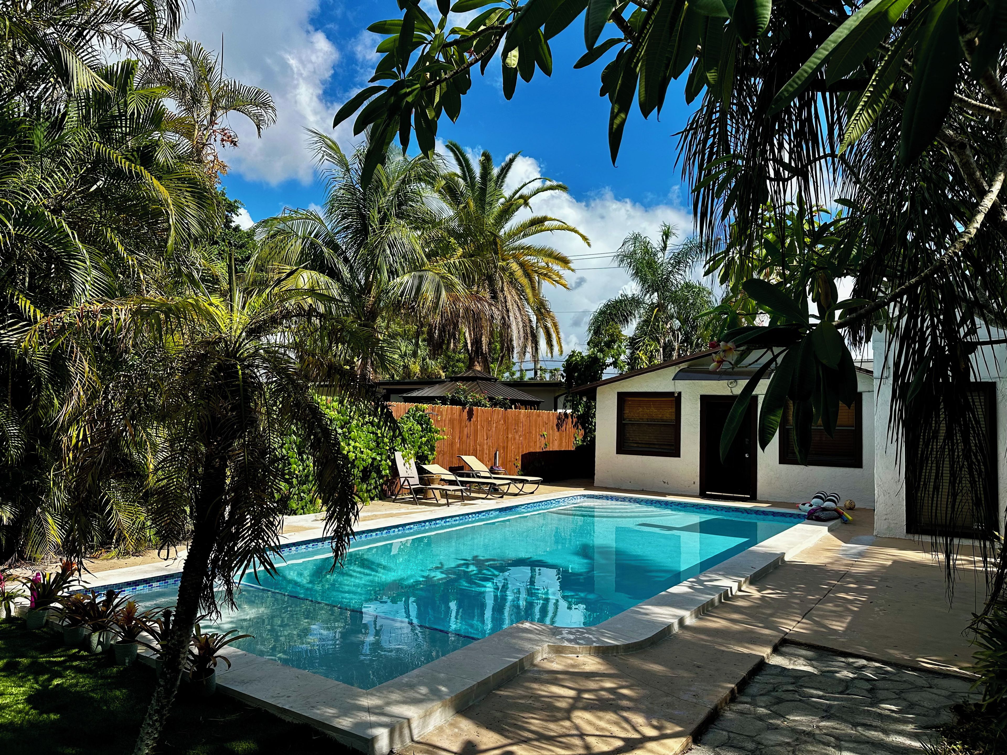 A swimming pool surrounded by lush tropical vegetation, a wooden deck, and a modern house with a sloped roof.