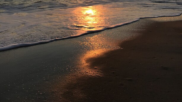 Ein Strand bei Sonnenuntergang, die Sonne spiegelt sich im Wasser.