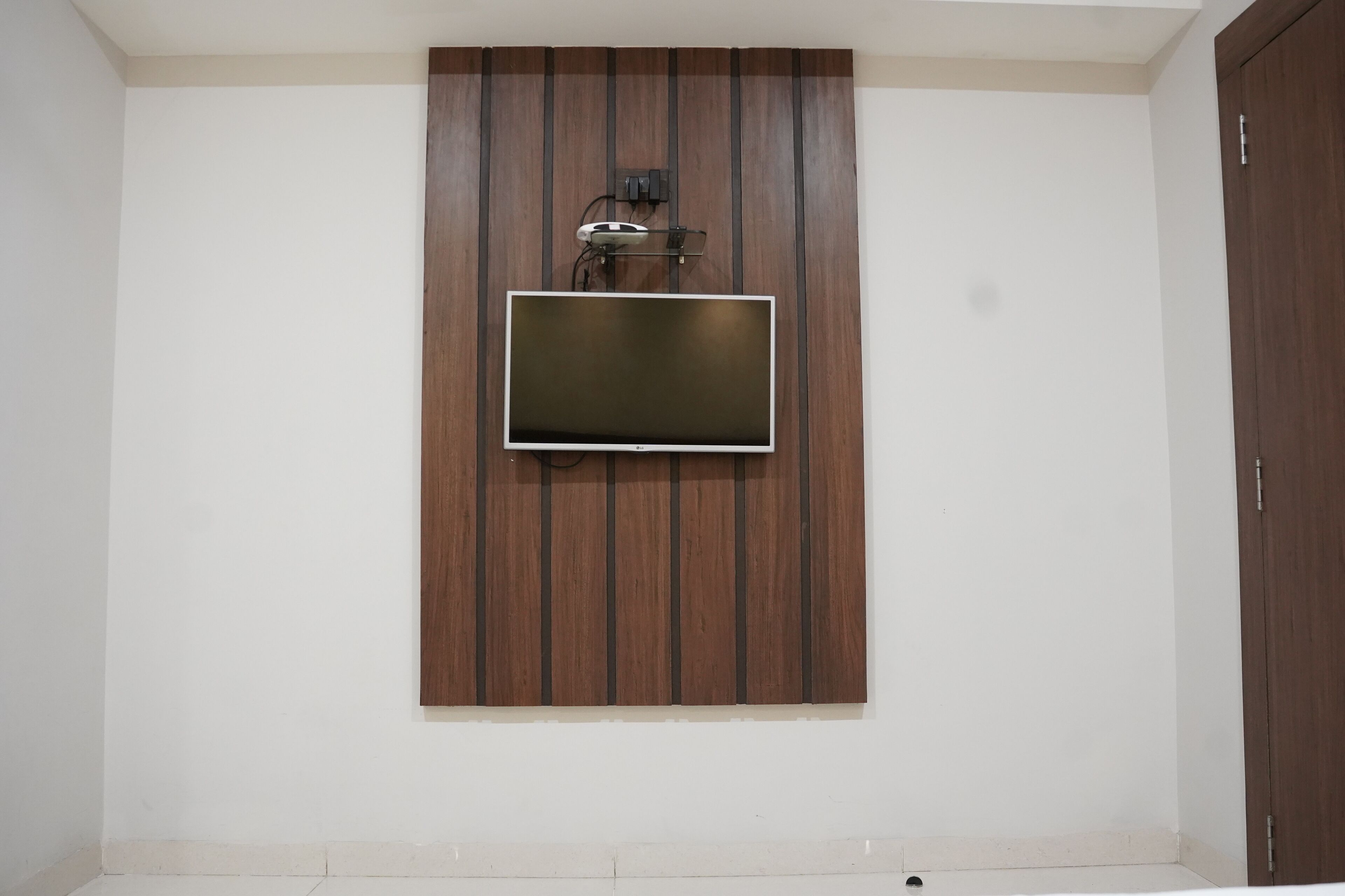 Une pièce avec un téléviseur à écran plat fixé au mur, qui est recouvert de panneaux en bois. Une porte se trouve sur le côté droit, et un interrupteur est placé sur le mur.