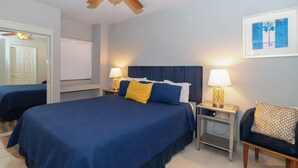 A hotel room with a large bed, two bedside tables with lamps, a ceiling fan, a framed picture on the wall, and a door leading to another room.