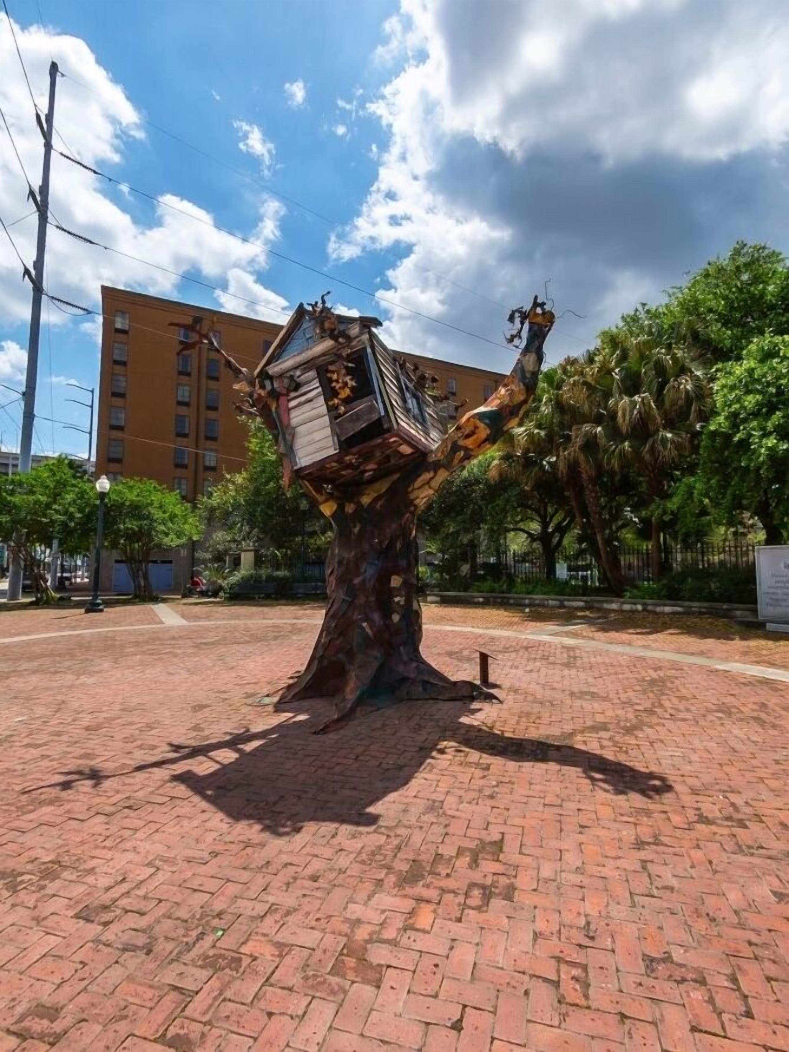 Uma escultura de árvore com uma casa em seus galhos, em uma área pavimentada com um edifício ao fundo.
