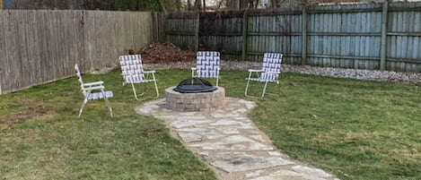 A backyard with a stone pathway leading to a fire pit surrounded by four patterned chairs.