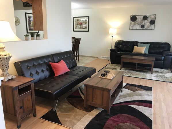 Ein Wohnzimmer mit einem schwarzen Ledersofa, einem Holztisch und einem Teppich. Zwei Lampen, eine Uhr und gerahmte Bilder schmücken die Wände.