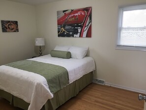 Ein ordentlich bezogenes Bett mit einer grün-weißen Tagesdecke, einer Lampe und einem an der Wand hängenden Bild von einem roten Fahrzeug.