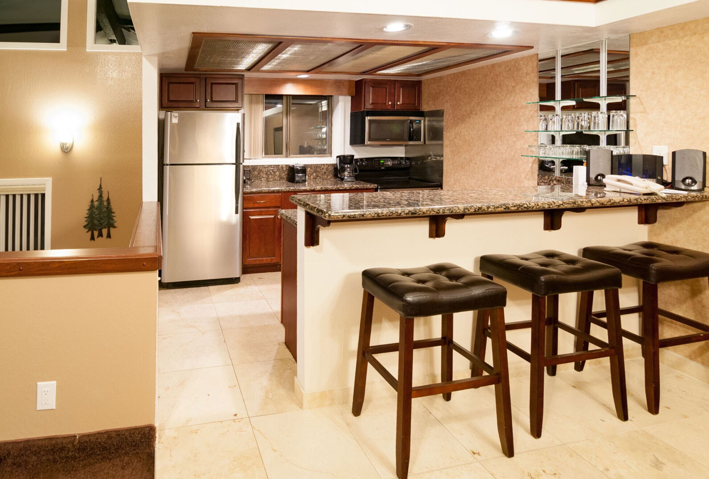 Cocina equipada con refrigerador, microondas y estufa. Hay tres taburetes de bar y una repisa de vidrio con vasos.