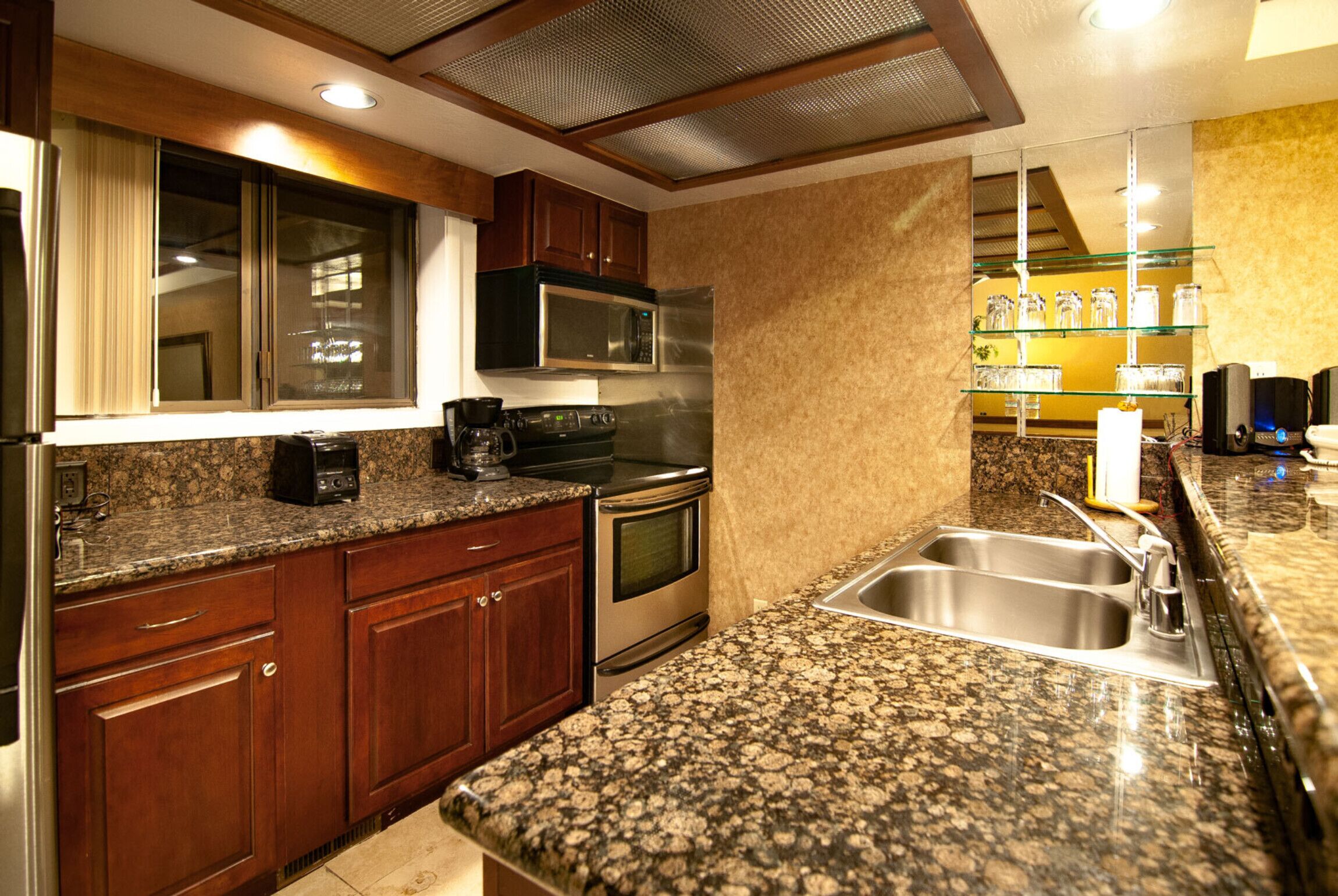 Una cocina con encimeras de granito, electrodomésticos de acero inoxidable, armarios de madera y una ventana con persianas.