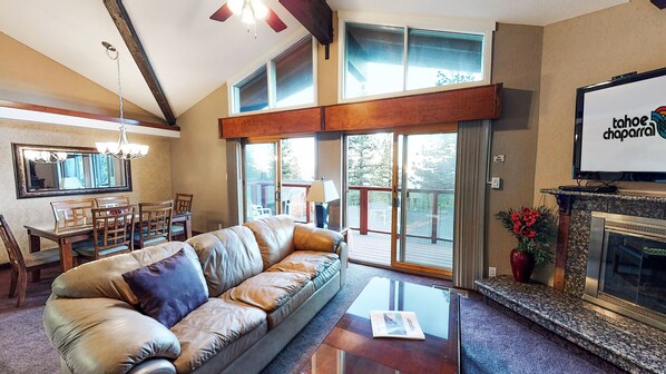 Amplia sala de estar con sofá de cuero, mesa de centro de vidrio y chimenea. Hay un comedor con mesa y sillas de madera, y ventanas grandes con vistas al exterior.