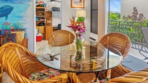 A round glass table surrounded by wicker chairs, a bookshelf, and a painting of a whale on the wall. - Kona Condo- Family Friendly- Fall Sale! (Kailua-Kona)