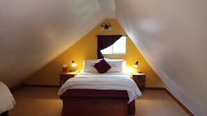 A bedroom with a sloped ceiling, a bed with white bedding and a red pillow, two bedside lamps, a nightstand, and a window with curtains. - Rustic luxury away from it all (Cloudcroft)