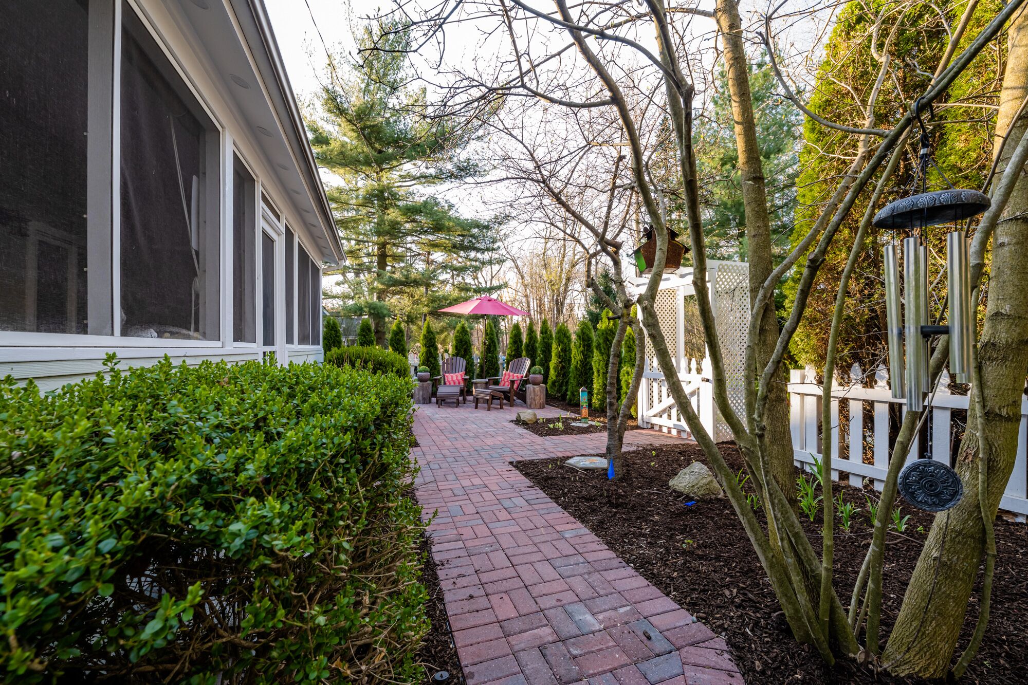 Un chemin pavé de briques mène à un espace patio aménagé avec du mobilier d’extérieur et délimité par une clôture blanche en bois.