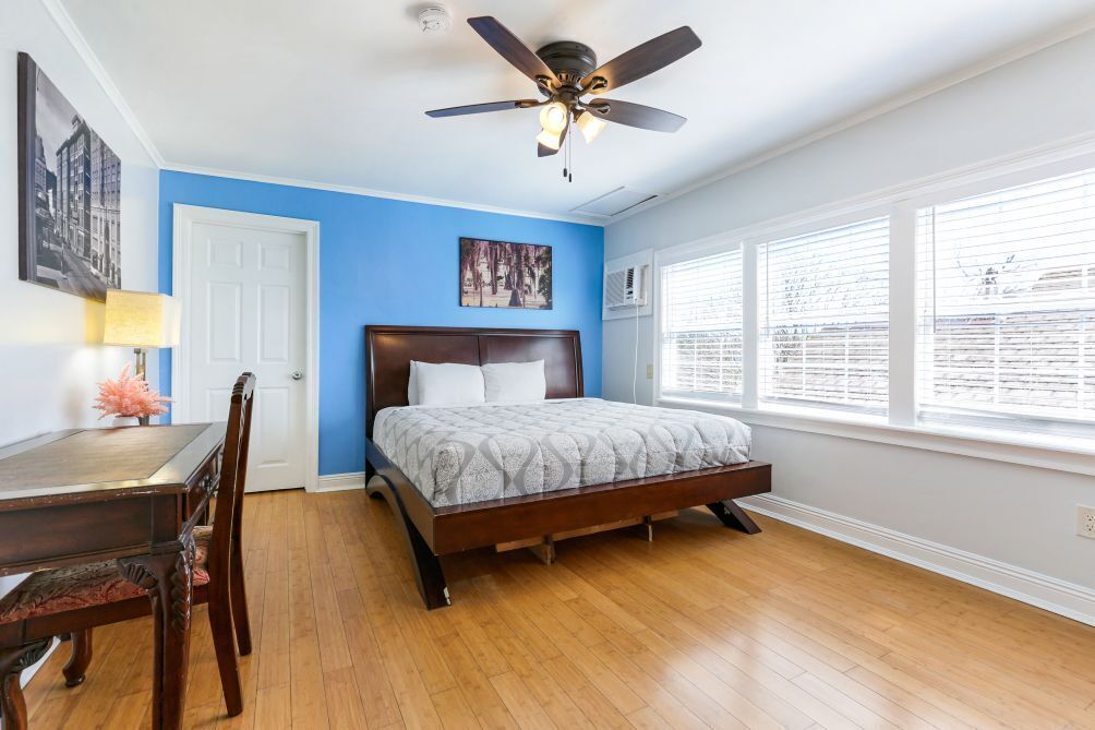A bedroom with a wooden floor, a bed with a headboard, a ceiling fan, and a window with blinds.