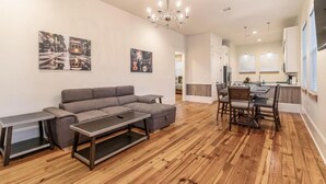 A modern living room with a sectional sofa, a dining table with chairs, wooden flooring, and a kitchen area in the background.