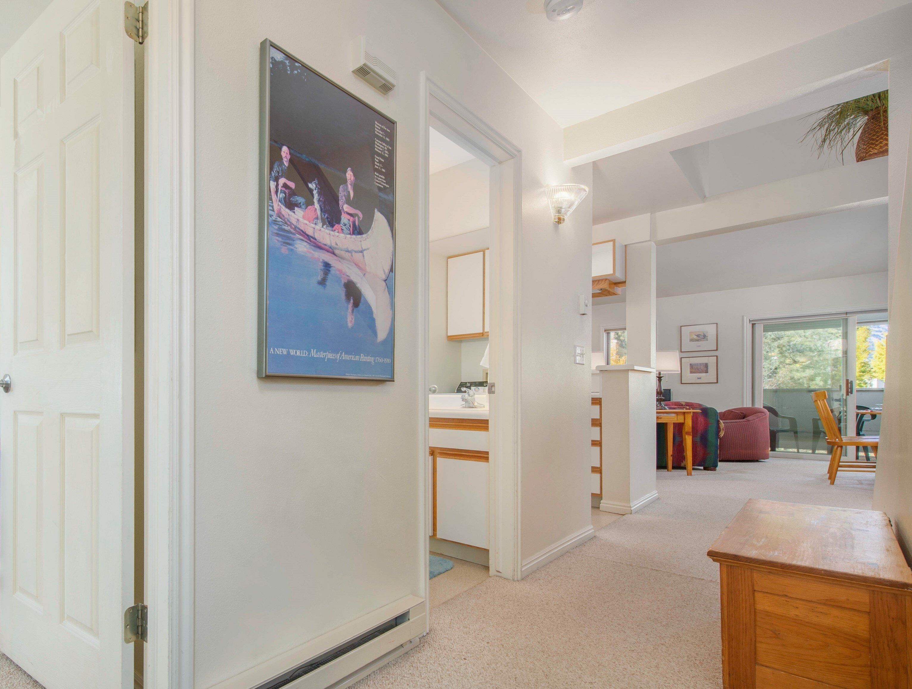 A hallway leading to a dining area with a poster on the wall, a kitchen area, and a view of the outdoors.
