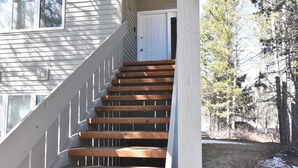 Un escalier en bois mène à une porte blanche, qui est entourée d’arbres et s’ouvre sur un ciel dégagé.