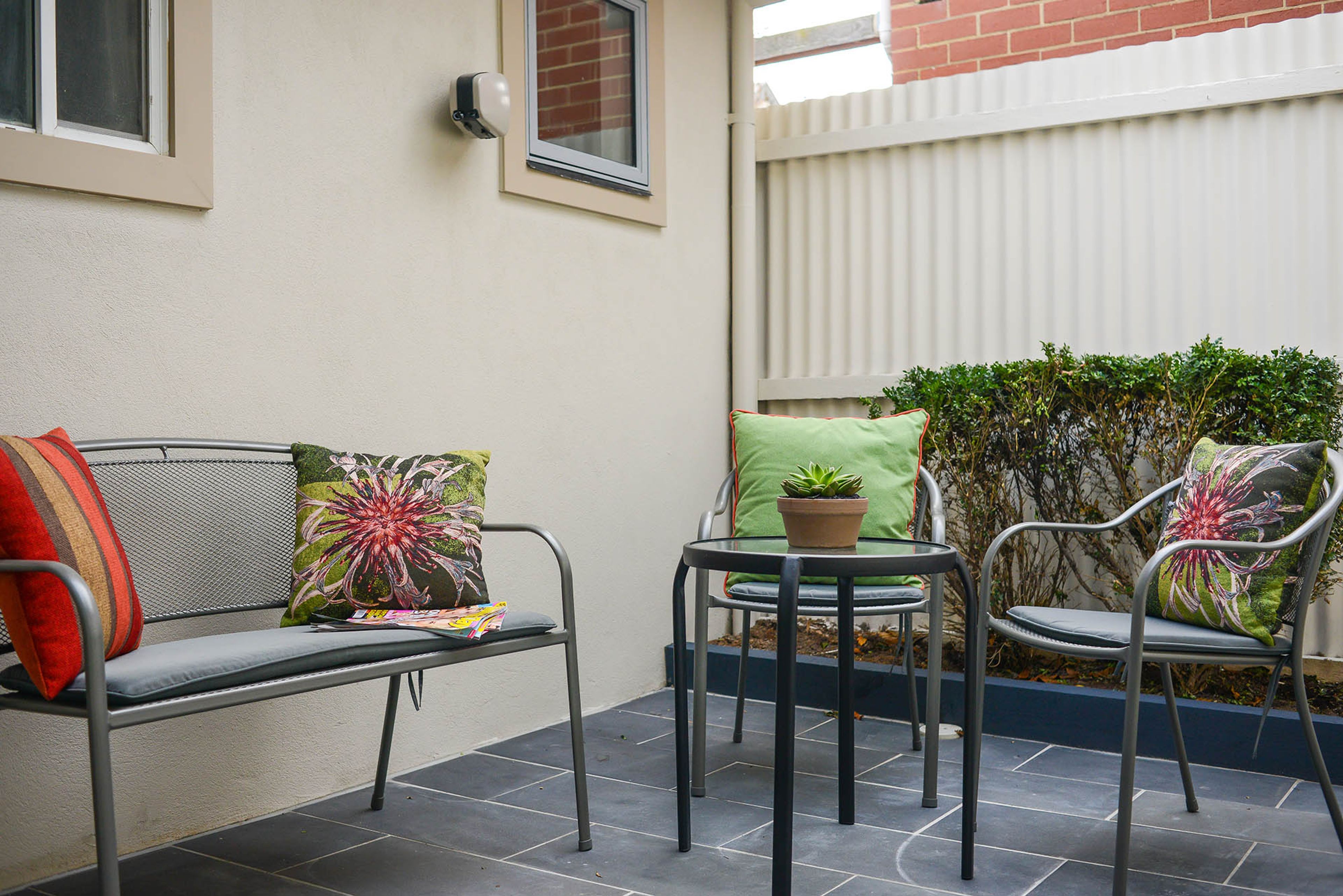 A patio with a metal bench, a small round table, and two chairs, each with a cushion.