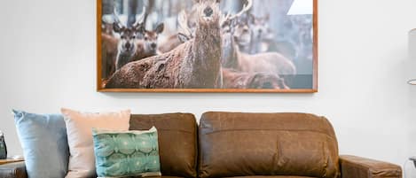Un sofá de cuero marrón con dos almohadones, uno liso y otro con estampado verde, en una habitación con una fotografía grande de un ciervo en la pared.