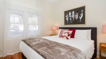 A bedroom with a large bed, a window with sheer curtains, a wall-mounted artwork, and a bedside table with a lamp.