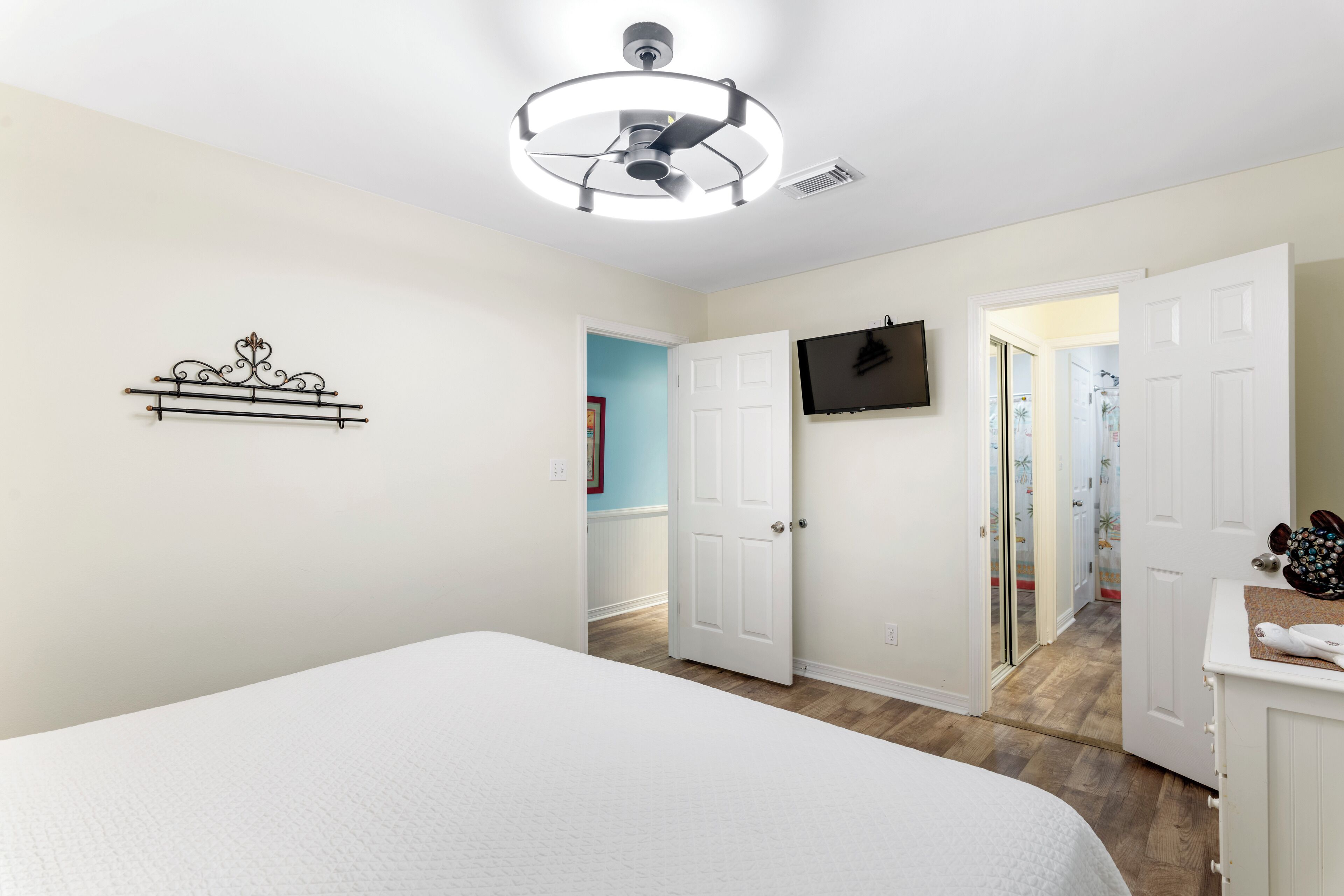 A bedroom with a bed, a ceiling fan, a mounted TV, and a door leading to another room.