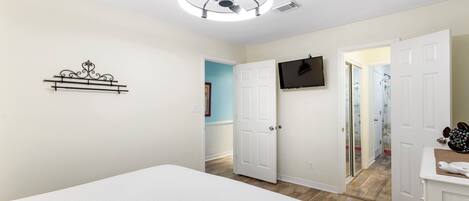 A bedroom with a bed, a ceiling fan, a mounted TV, and a door leading to another room.