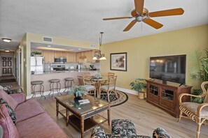 A living room with a sofa, coffee table, and chairs, a dining area with a table and chairs, a kitchen with appliances, and a ceiling fan.