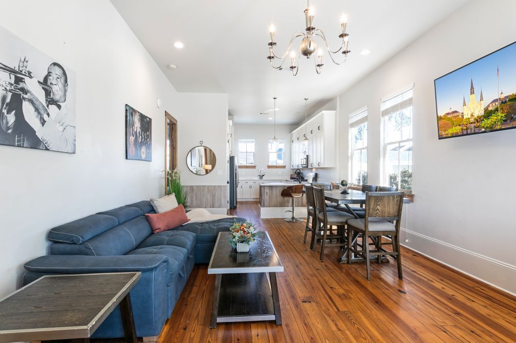 A modern living room with a blue sofa, a dining table with chairs, a chandelier, and a kitchen in the background.