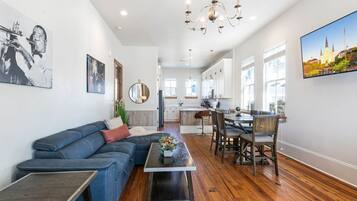 A modern living room with a blue sofa, a dining table with chairs, a chandelier, and a kitchen in the background.