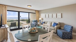 Ein geräumiges Wohnzimmer mit einem großen Glasfenster, das auf einen Balkon führt, einem runden Glastisch und einer Sofagarnitur.