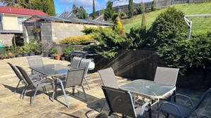 Un patio al aire libre con una mesa de vidrio y sillas de metal, rodeado de plantas en macetas y un muro de piedra.